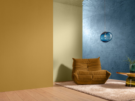 Ein gemütlicher Raum mit zwei Wänden in unterschiedlichen Farben – eine gelbe und eine blaue Wand. In der Ecke steht ein brauner Sessel. Auf dem Boden liegt ein weicher, heller Teppich. Neben dem Sessel steht ein runder Tisch aus Holz mit einer modernen, grünen Vase, die eine Pflanze enthält. An der Decke hängt eine stilvolle, transparente Lampe.