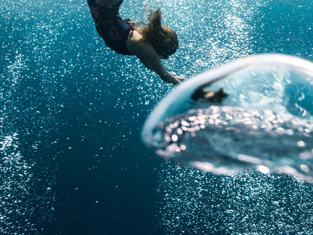 Eine Unterwasseraufnahme, die einen Menschen zeigt, der in einem blauen Wasser schwimmt. Die Person ist teilweise untergetaucht und zieht ihre Arme nach vorne. Im Vordergrund sieht man eine große Wasserblase.