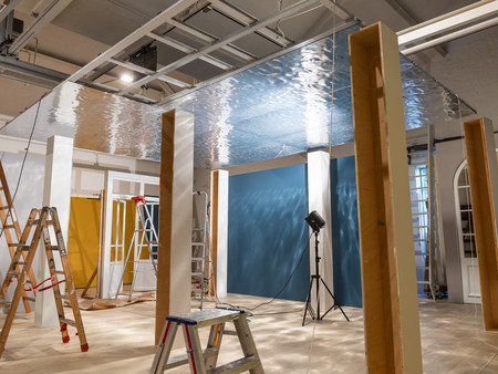 Ein Raum mit Leitergerüsten, in dem ein reflektierendes, metallisches Panel an der Decke angebracht ist. Es gibt mehrere Leitern im Raum, und im Hintergrund sind eine gelbe Wand und eine blaue Wand sichtbar. 