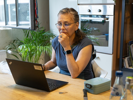 Eine Frau mit blonden, schulterlangen Haaren sitzt an einem Holztisch und schaut auf ihren Laptop. Sie trägt eine graue Oberbekleidung und eine Armbanduhr. Ihr Hand ist am Kinn, während sie nachdenklich aussieht