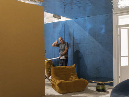 Ein Mann steht in einem Raum und zeigt "Dauem hoch". Im Hintergrund sind eine blaue Wand und gepolsterte Möbel in warmen Farben zu sehen. 