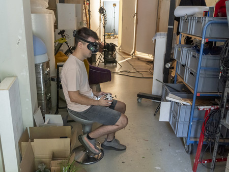 Ein junger Mann sitzt auf einem Stuhl in einem Raum mit wenig Licht, während er ein VR-Headset trägt. Er hält einen Controller in der Hand.