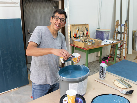 Ein Mann in einem grauen T-Shirt mischt Farbe in einen großen, grauen Eimer. Er lächelt und hält eine Tasse in der Hand. Im Hintergrund sind verschiedene Werkzeuge und Materialien sichtbar, die für Malerarbeiten verwendet werden.