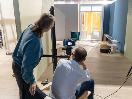 Eine Frau mit langen, lockigen Haaren in einem blauen Sweatshirt steht hinter einem Mann, der auf einem Stuhl sitzt und auf einen Bildschirm schaut.