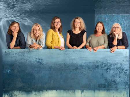Eine Gruppe von sechs Frauen steht hinter einem großen, blauen Gemälde, das eine abstrakte, hellblaue und grüne Farbverlaufgestaltung zeigt.
