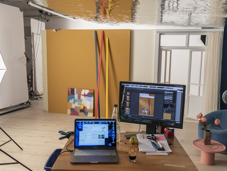 Ein kreatives Studio mit einem Tisch, auf dem ein Laptop und ein Monitor stehen. Im Hintergrund sind verschiedene kreative Materialien sichtbar, darunter farbige Stangen und eine große gelbe Wand. Eine Kamera- und Beleuchtungsausrüstung ist ebenfalls im Raum.
