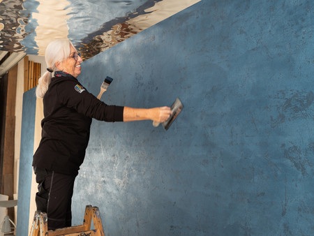 Eine Frau steht auf einer Leiter und führt eine Kreativtechnik an der blauen Wand aus. Sie hat graue Haare, trägt eine Brille und arbeitet mit einem Lächeln im Gesicht.