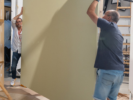 Zwei Personen heben eine große Wandplatte in einem Atelier. Die Umgebung zeigt Baugerüste und andere Materialien.