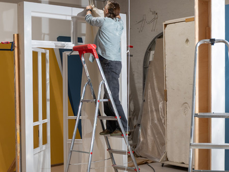 Eine Person steht auf einer Leiter und arbeitet an einer Decke in einem Raum. Sie trägt eine lockere Hemd und ist mit einem Bauprojekt beschäftigt. Um sie herum sind verschiedene Materialien und Werkzeuge, darunter eine weitere Leiter und Abdeckungen auf dem Boden. 