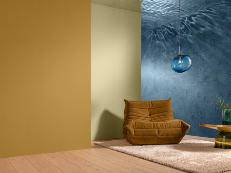 Ein gemütlicher Raum mit zwei Wänden in unterschiedlichen Farben – eine gelbe und eine blaue Wand. In der Ecke steht ein brauner Sessel. Auf dem Boden liegt ein weicher, heller Teppich. Neben dem Sessel steht ein runder Tisch aus Holz mit einer modernen, grünen Vase, die eine Pflanze enthält. An der Decke hängt eine stilvolle, transparente Lampe.