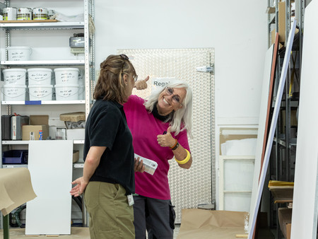 Zwei Frauen tauschen sich in einem Atelier aus, wobei eine Frau "Daumen hoch" zeigt. Im Hintergrund Regale mit zahlreichen, beschrifteten Behältern für Farben und Materialien.