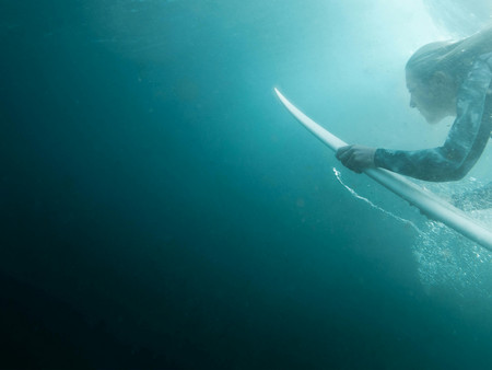 Ein Surfer taucht unter Wasser mit einem Surfbrett. Die Lichtverhältnisse sind gedämpft, und das Wasser hat eine tiefblaue Farbe.