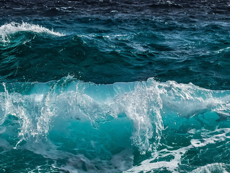 Eine Nahaufnahme von tobenden Wellen im Ozean. Die Wellen zeigen verschiedene Blau- und Grüntöne, während sie an der Oberfläche schäumen und spritzen. 
