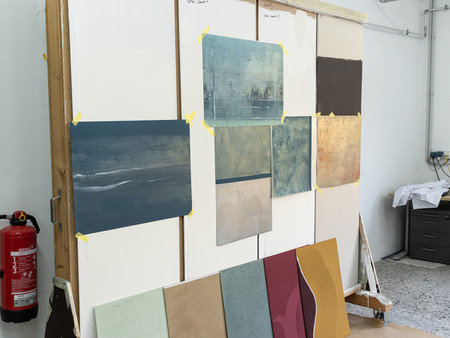 Ein Kunststudio, in dem mehrere Gemälde an einer Wand hängen. Die Bilder sind mit Klebeband befestigt und zeigen verschiedene abstrakte Landschaften in unterschiedlichen Farbnuancen. Im Vordergrund liegen weitere Gemälde auf dem Boden.