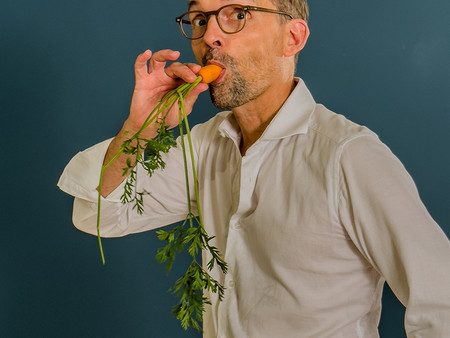 Ein Mann mit Brille hält eine große Karotte mit frischem Grün in der Hand und beißt leicht daran. Er trägt ein weißes Hemd und steht vor einem blauen Hintergrund.