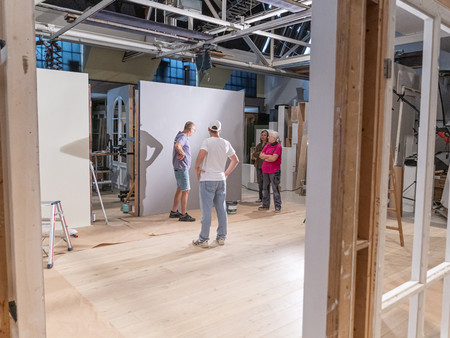 Ein Team von vier Personen steht in einem großen, hellen Raum, der mit weißen Wänden und einem hellen Holzboden ausgestattet ist. Es gibt Leitern und Malutensilien, und die Personen scheinen an einem Projekt zu arbeiten, während sie sich miteinander unterhalten. 