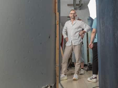Ein Mann mit Brille und einem hellen Hemd steht selbstbewusst mit den Händen in den Hüften in einer Studio-Umgebung. 