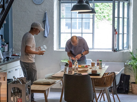 Ein Mann in einer Küche, der eine Glasflasche hält, während ein anderer Mann am Tisch steht und Zutaten zum Essen vorbereitet. 