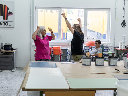 Drei Personen in einem hellen, kreativen Raum. Zwei Personen, eine Frau mit grauen Haaren und ein Mann, heben ihre Arme in die Luft und scheinen sich zu freuen. Im Hintergrund sitzt ein weiterer Mann an einem Tisch, während verschiedene Malutensilien und Farben auf der Arbeitsfläche verteilt sind.