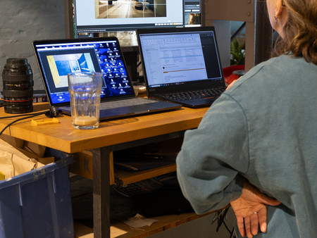 Eine Person steht vor einem Tisch mit zwei Laptops und einem Monitor. Auf dem Tisch befindet sich ein Glas und eine Kameralinse.