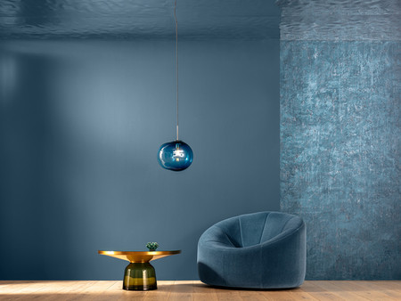 Ein stilvoll eingerichteter Raum mit einer blauen Wand und einer strukturierten Farbanpassung. In der Mitte steht ein runder, goldfarbener Tisch mit einer kleinen Pflanze darauf. Zu sehen ist auch ein bequemer, rund geformter Sessel in einem sanften Blauton. Über dem Tisch hängt eine moderne, runde Lampe in Blau. 