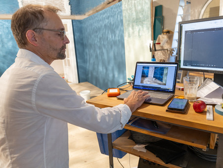 Ein Mann sitzt an einem Tisch und arbeitet an einem Laptop. Er trägt ein weißes Hemd und hat eine Brille auf. 