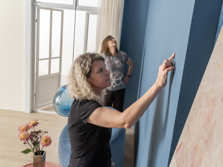 Das Bild zeigt zwei Frauen in einem hellen Raum. Eine Frau mit lockigem, blondem Haar steht im Vordergrund und streicht mit ihrem Finger über eine blaue Wand. Die andere Frau steht im Hintergrund und betrachtet die Wand. 