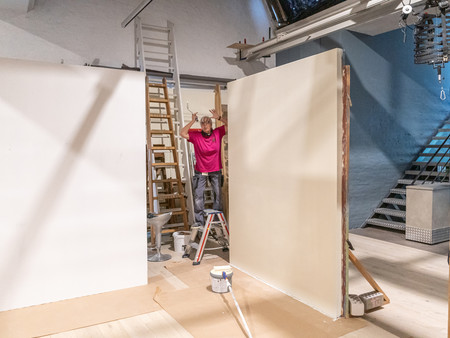 Eine Frau in einem pinken Shirt hält die Hände an den Kopf. Im Hintergrund sind unbemalte Wände zu sehen, und der Raum ist teilweise mit Treppen und Malerutensilien ausgestattet. Der Boden ist mit Malerpapier ausgelegt.