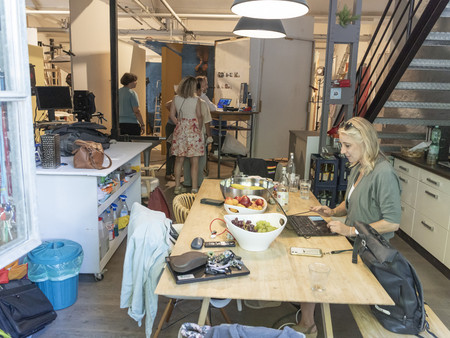In einem kreativen Arbeitsraum sitzen mehrere Personen. Eine Frau mit blonden Haaren arbeitet an einem Laptop auf einem Holztisch. Im Hintergrund sind einige weitere Menschen zu sehen, die sich unterhalten oder an einem Projekt arbeiten. 