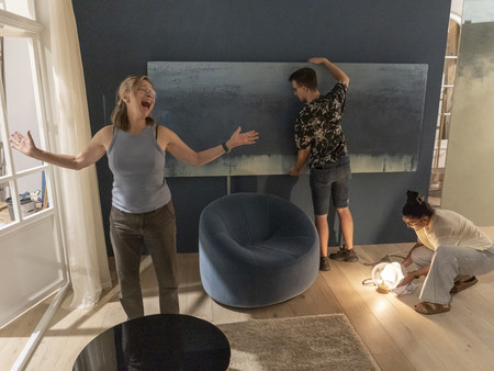 Eine Frau steht lachend in einem Raum mit blauen Wänden. Im Hintergrund sind zwei Personen sichtbar: Ein Mann, der einen großes Bild an die Wand hängt und eine Frau, die am Boden mit einer Lampe arbeitet. 