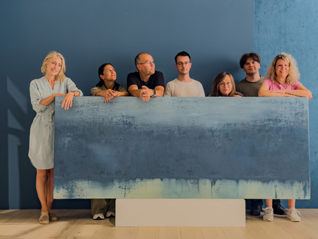 Eine Gruppe von sieben Menschen steht hinter einem großen, blauen Bild, das eine abstrakte, hellblaue und grüne Farbverlaufgestaltung zeigt. 