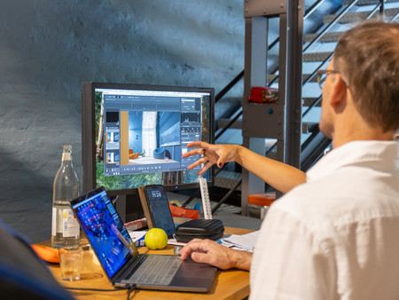 Eine Person sitzt an einem Tisch und arbeitet am Computer. Auf dem Bildschirm ist eine Bearbeitungssoftware für Bilder oder Design zu sehen. Die Person zeigt mit der Hand auf den Bildschirm.