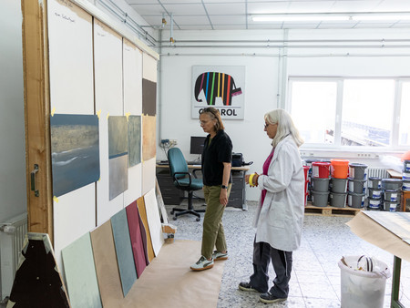 Zwei Frauen in einem Atelier. Eine Frau mit langen Haare steht an einer Wand mit verschiedenen Kunstwerken oder Farbmustern, während die andere Person, die einen weißen Kittel trägt, in der Nähe steht und sie betrachtet. Im Hintergrund sind Stühle und Farbdosen zu sehen.