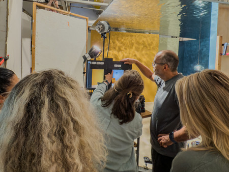 Eine Gruppe von Menschen arbeitet in einem Studio, umgeben von professionellen Foto- und Videoausrüstungen. Eine Person zeigt auf einen Bildschirm, während andere aufmerksam zuschauen.