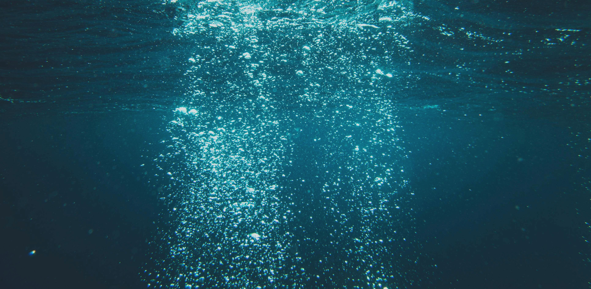 Unterwasseraufnahme, die eine Reihe von Luftblasen zeigt, die im blauen Wasser aufsteigen, mit Lichtreflexionen, die die glitzernde Oberfläche betonen.